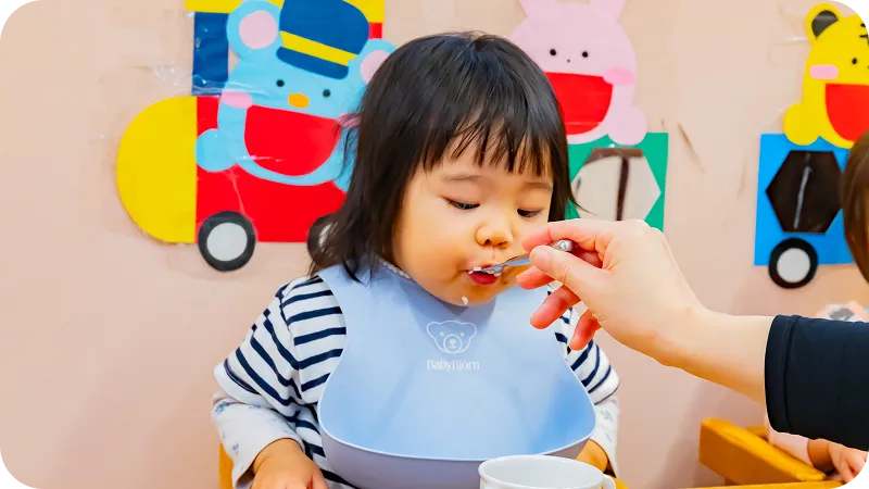 保育士に介助されながらスプーンで離乳食を食べる乳児の様子